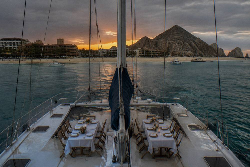 A catamaran deck with tables set for dinner as the sky turns orange over the water