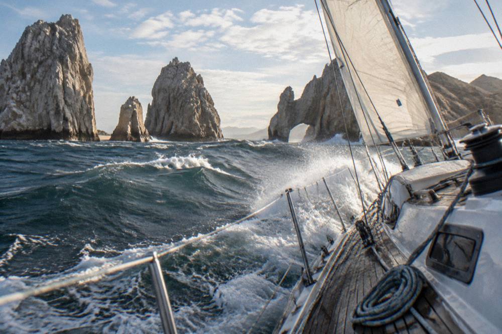 A sailing yacht underway near rock formations at the tip of the Baja peninsula in late afternoon light