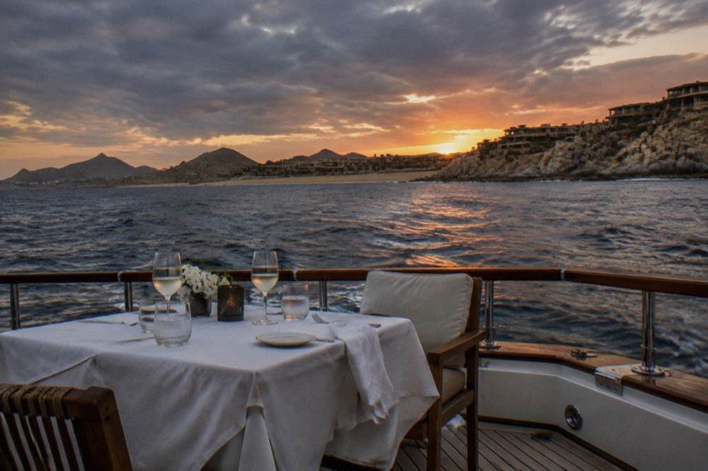 An evening table for two on a boat deck with glasses raised toward an orange horizon