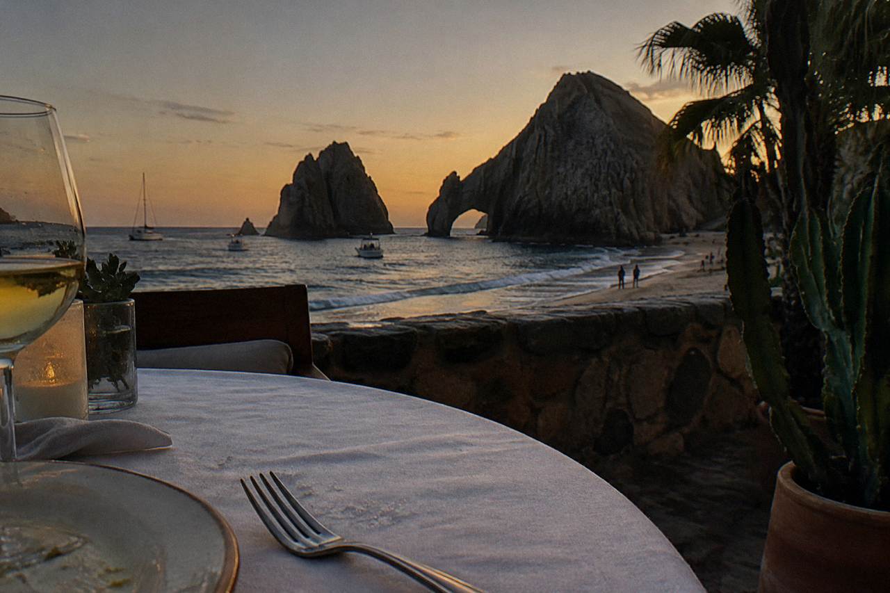 A dinner table set on the deck of a boat facing the Cabo San Lucas Arch at dusk