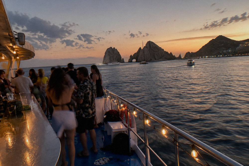 A crowded open deck of a boat at dusk with string lights and a bar counter along one side.