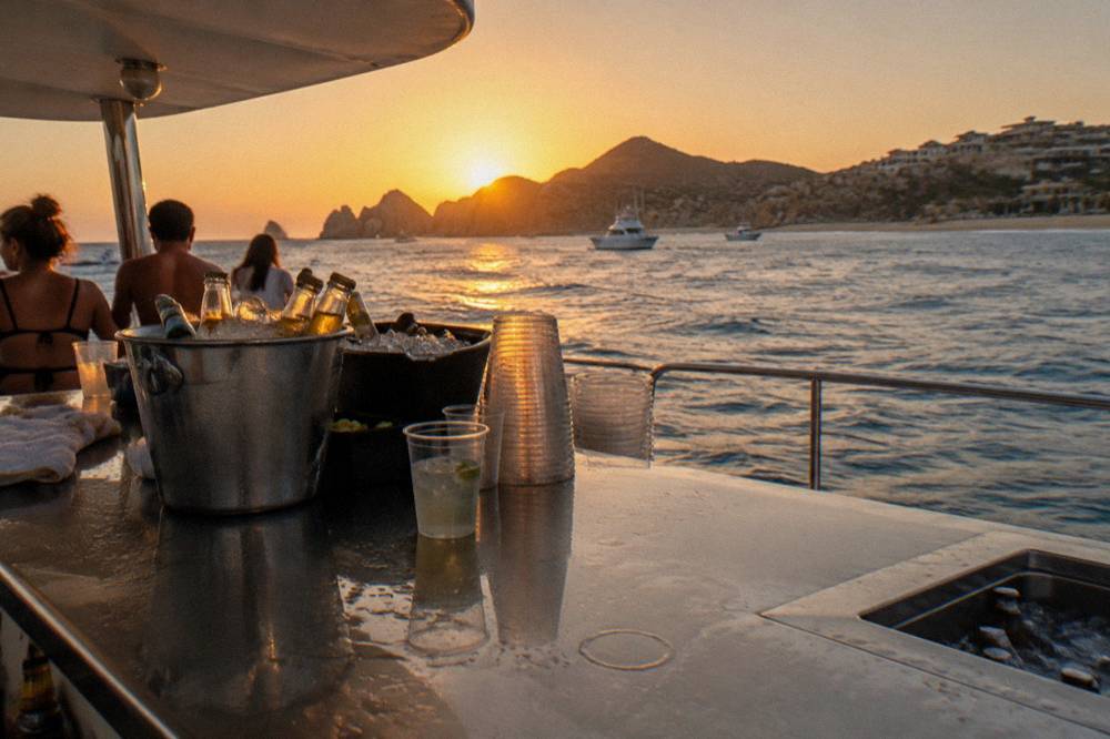 A boat bar counter with plastic cups, ice buckets and bottles set against an ocean horizon at dusk.