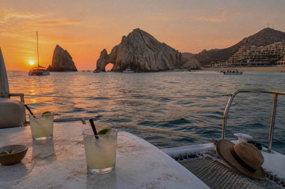 A wide catamaran deck under an orange sunset sky with drinks on a table and rock formations in the distance.