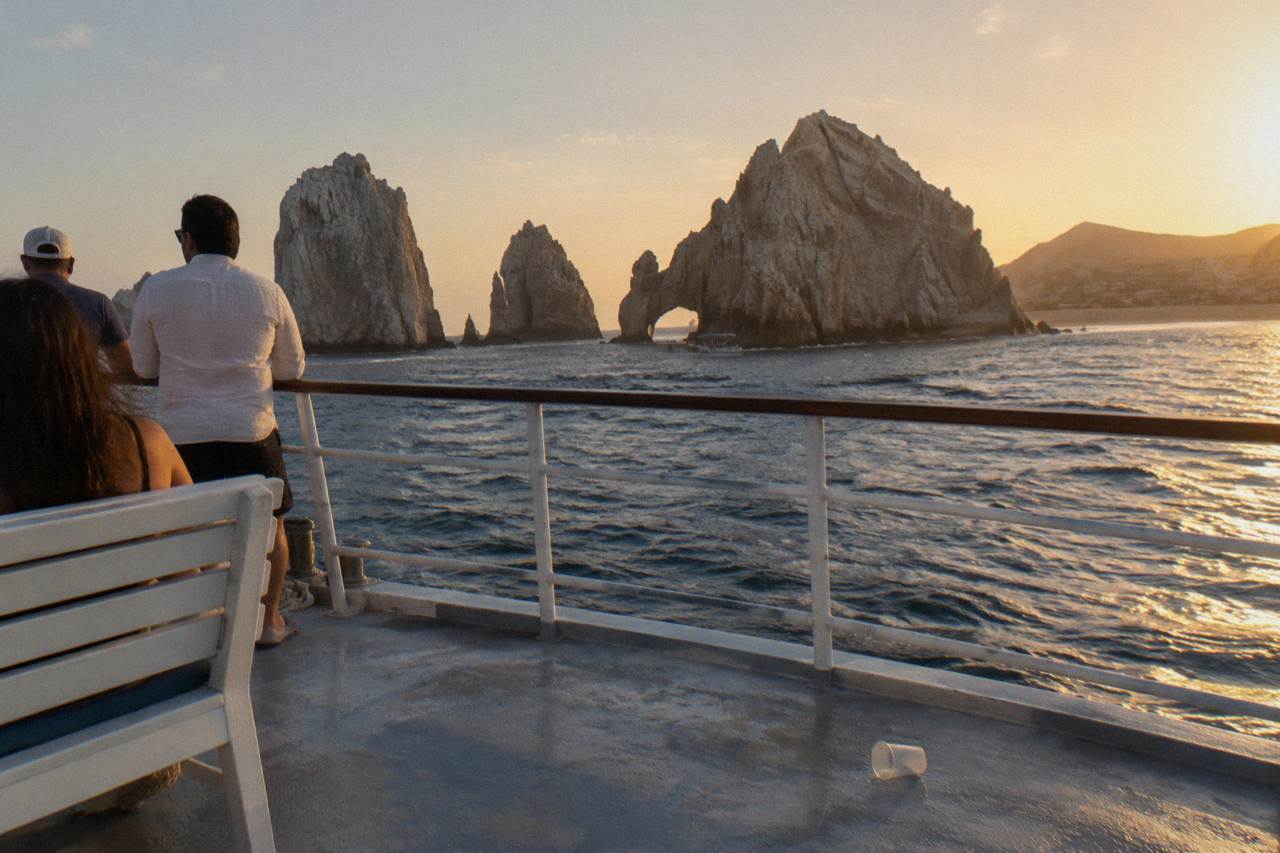 A party boat underway near rock formations at the tip of the Baja peninsula under a golden evening sky.