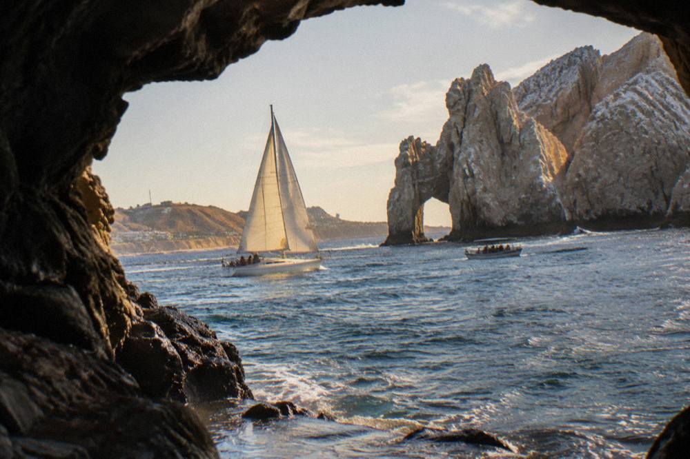 A sailing yacht with sails up cruising past warm-lit rocks near the Cabo marina