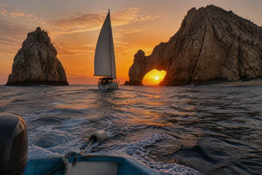 A sailboat under orange sunset sky near a stone arch