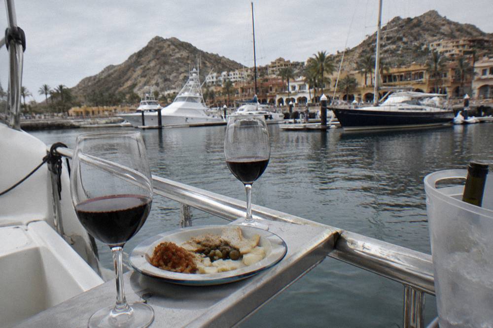 Glasses of wine and a plate of appetizers resting on a wooden deck rail with open water behind