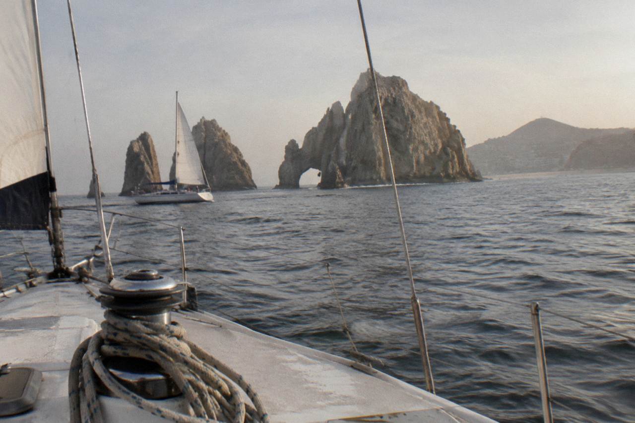 A sailboat under warm light gliding across calm water near rock formations at the tip of the Baja peninsula