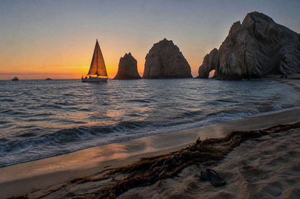 Sailboat under warm evening light on open water near dark shoreline rocks