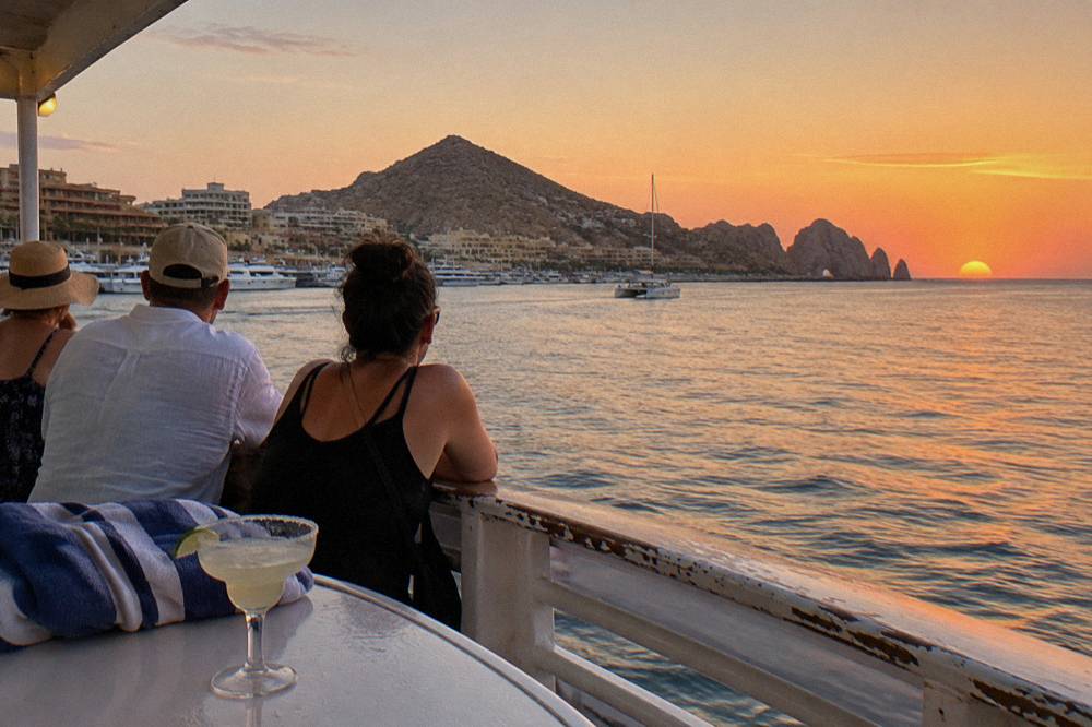 Passengers at the rail of a boat watching an orange sunset sky over calm water near Cabo San Lucas