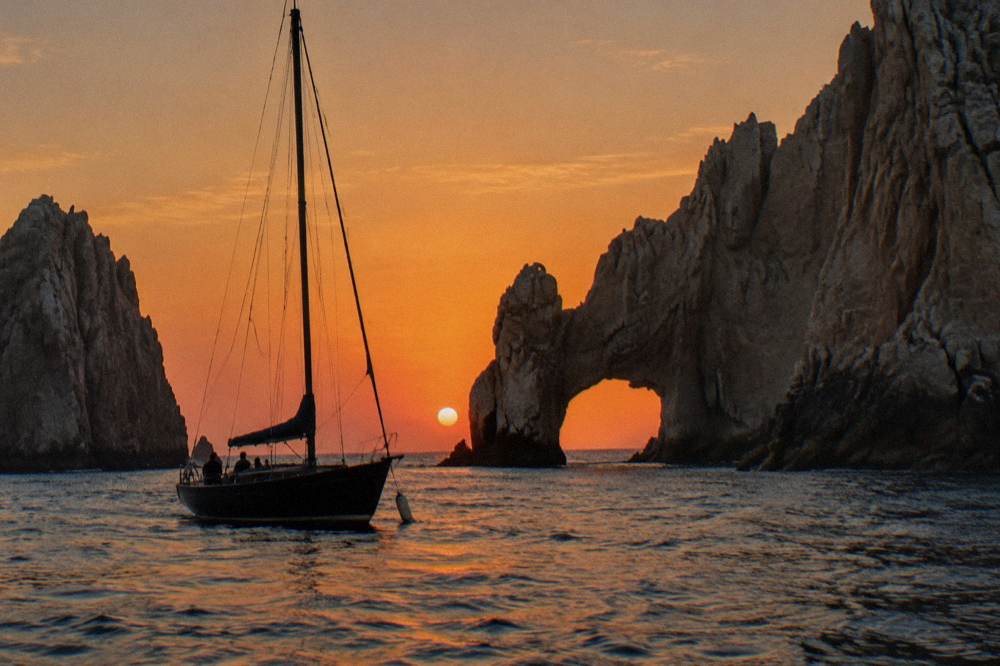 Sailboat with passengers silhouetted against an orange sky near a rock arch