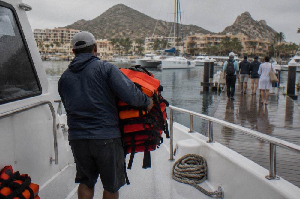 Crew member handing life vests to passengers at a marina dock beside a white tour boat