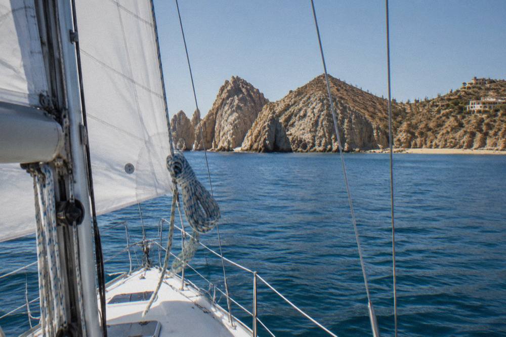 White sailing yacht underway with full sails on calm blue water near the Cabo San Lucas coastline