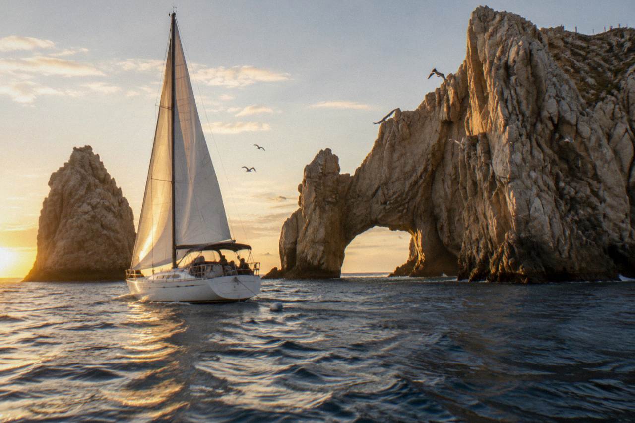 Sailboat with white sails raised passing the rock arch at Lands End near Cabo San Lucas at golden hour