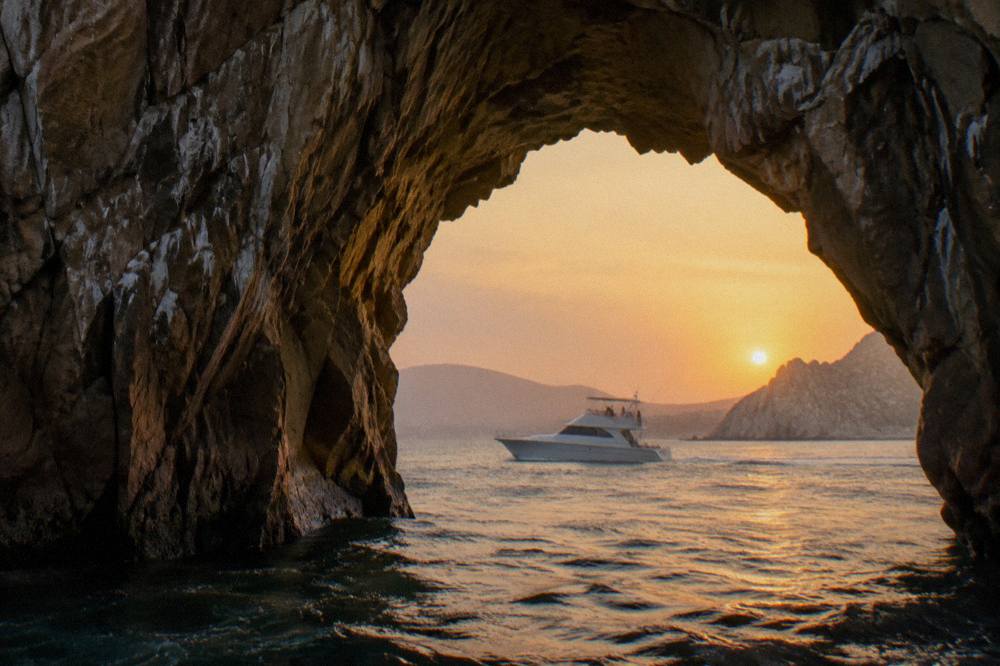 Sleek white yacht cruising past golden rock formations near the Cabo San Lucas arch at sunset