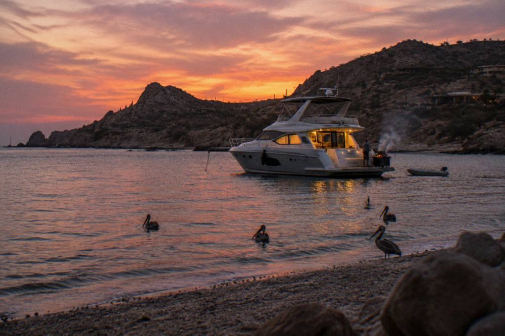 Private yacht at anchor under a pink and orange sky with dark silhouetted hills behind Cabo San Lucas
