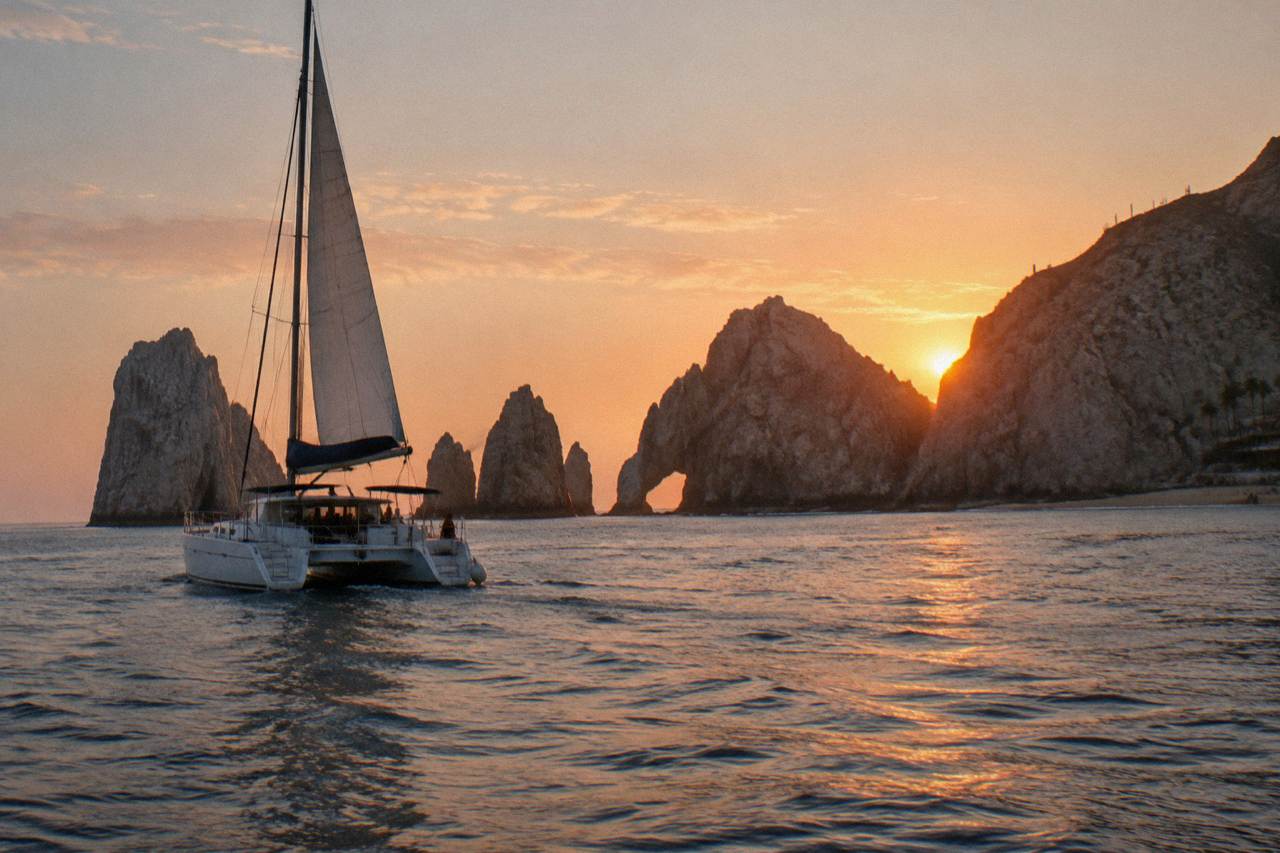 A catamaran under sail near rock formations at the end of the Cabo San Lucas coastline at sunset