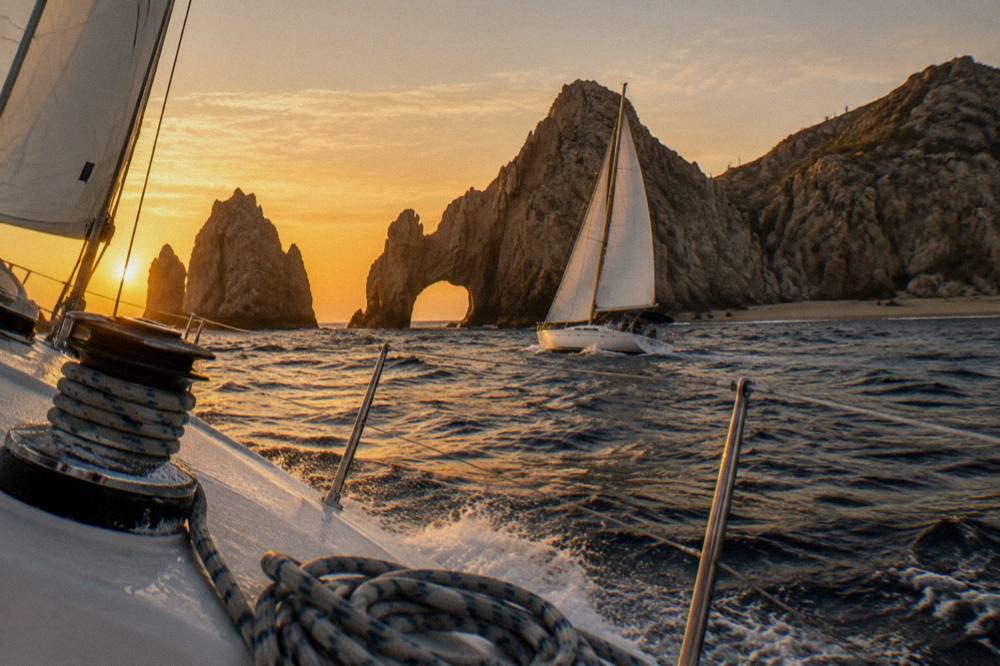 Sailboat heeling across golden water near a rocky arch formation at dusk