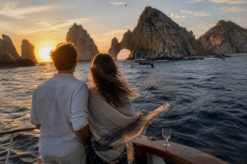 Couple standing at the stern of a yacht with the Cabo San Lucas Arch lit by sunset behind them