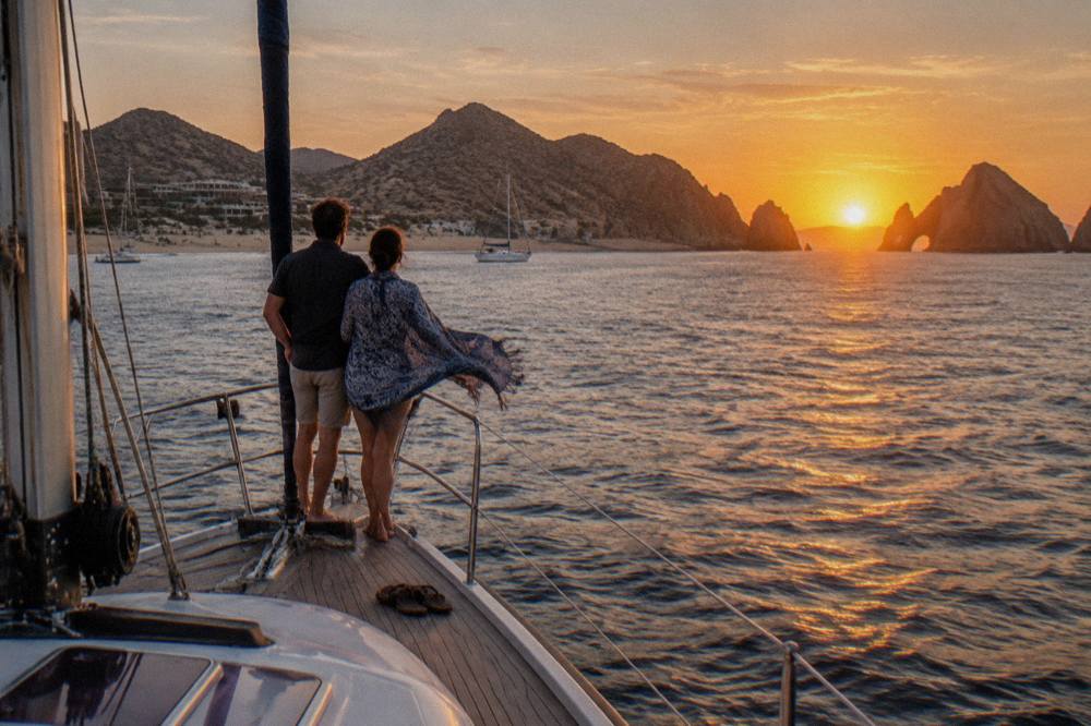 A couple at the railing of a sailboat with orange sunset light over open water