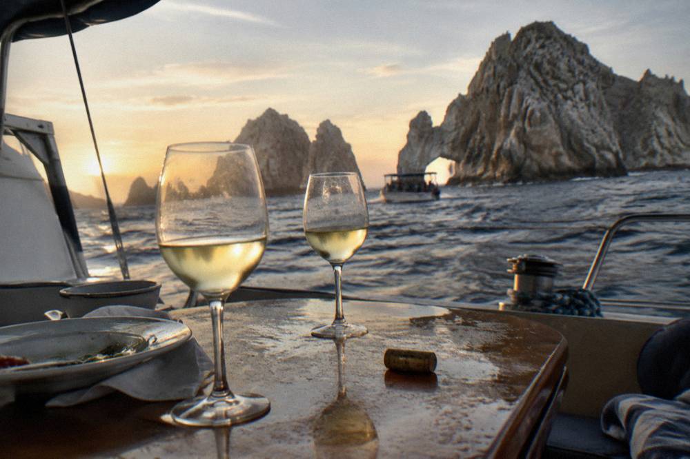 Two wine glasses on a boat table with the rock arch in the background at golden hour