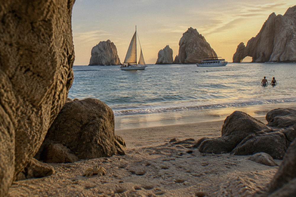 A sailing yacht passing El Arco at golden hour while a dinner boat sits at anchor nearby