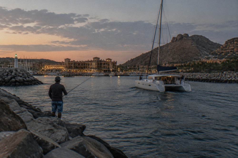 Catamaran with passengers on deck cruising past the Cabo San Lucas coast at dusk