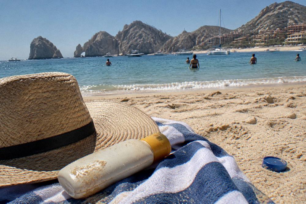 Beach towel, hat and sunscreen arranged on sand beside a calm bay at midday