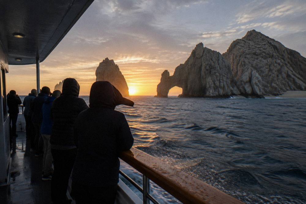 Passengers in windbreakers standing at a boat rail as the sun sets near a rock arch