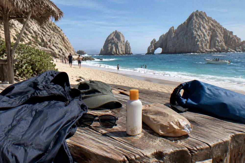 A flat-lay of a windbreaker, cap, sunglasses, sunscreen and a small dry bag on a wooden table